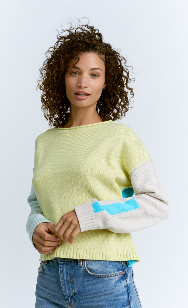 Front top half view of a woman wearing jeans and the lisa todd color crush sweater. This sweater is green/yellow in the front. It has drop shoulder long sleeves. The left sleeve is white while the right sleeve is pastel blue. The back of the sweater is sky blue.