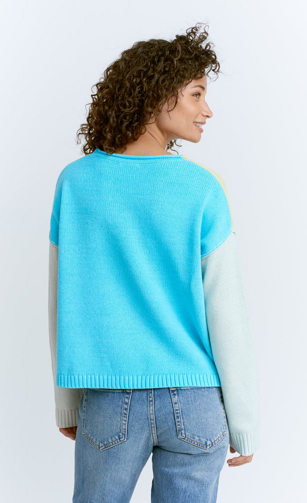 Back top half view of a woman wearing jeans and the lisa todd color crush sweater. This has drop shoulder long sleeves. The left sleeve is white while the right sleeve is pastel blue. The back of the sweater is sky blue.