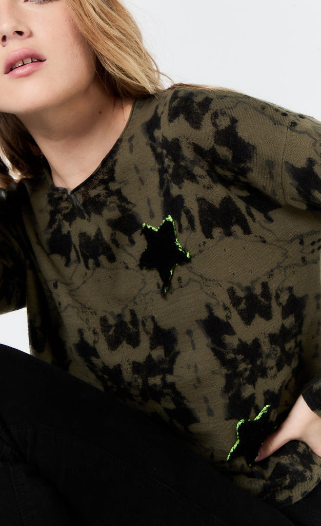 Front top half view of a woman wearing the lisa todd popstar top in the color rainforest (olive with black tie dye). This top has long sleeves, a round neck, and a front star shaped patch.