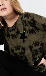 Load image into Gallery viewer, Front top half view of a woman wearing the lisa todd popstar top in the color rainforest (olive with black tie dye). This top has long sleeves, a round neck, and a front star shaped patch.
