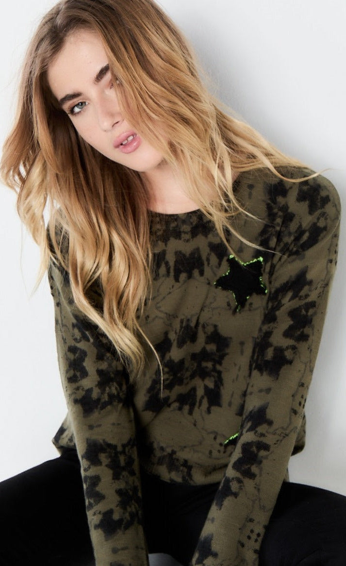 Front top half view of a woman wearing the lisa todd popstar top in the color rainforest (olive with black tie dye). This top has long sleeves, a round neck, and a front star shaped patch.