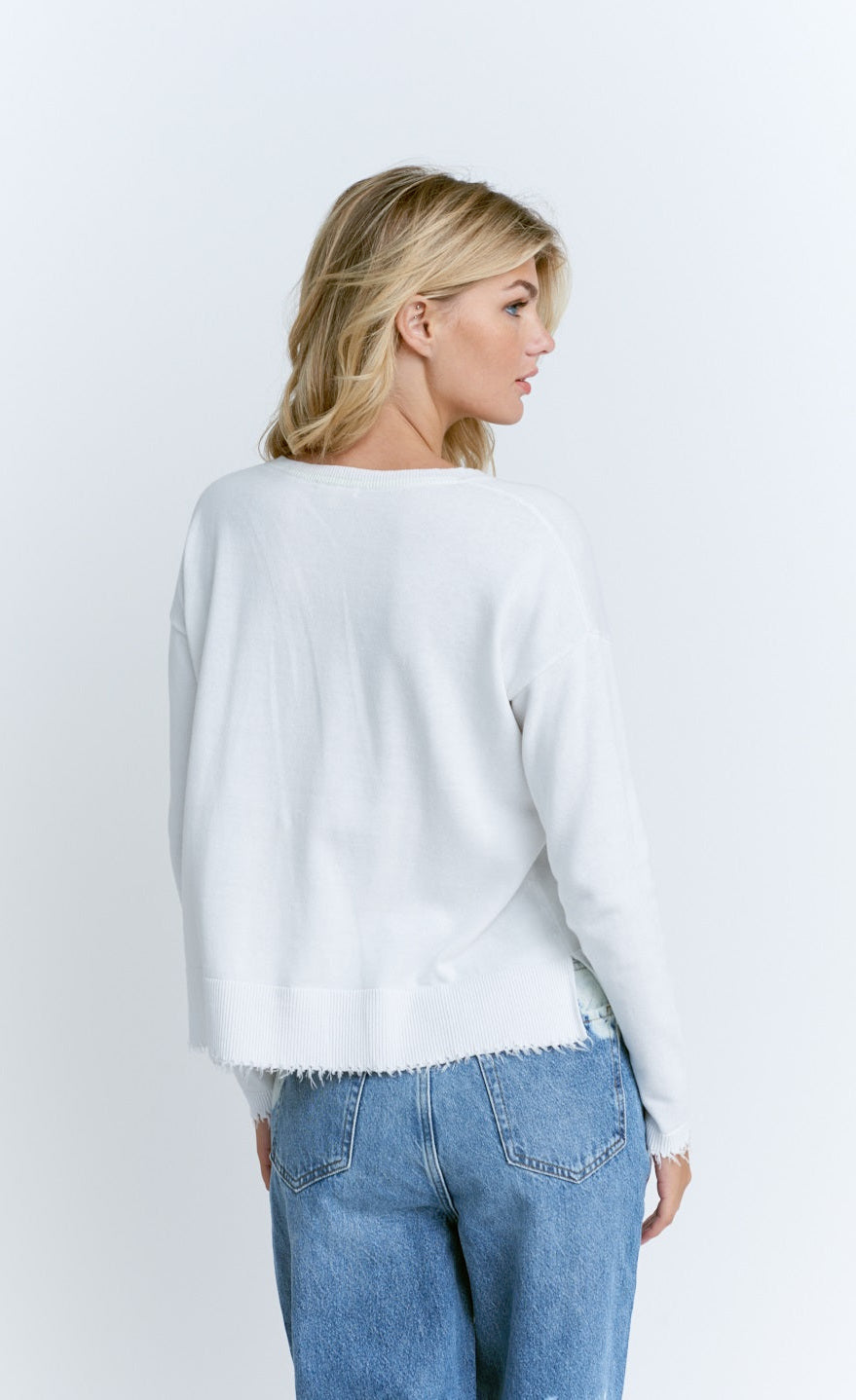 Back top half view of a woman wearing the lisa todd tainted love sweater. This sweater is white with a frayed hem.