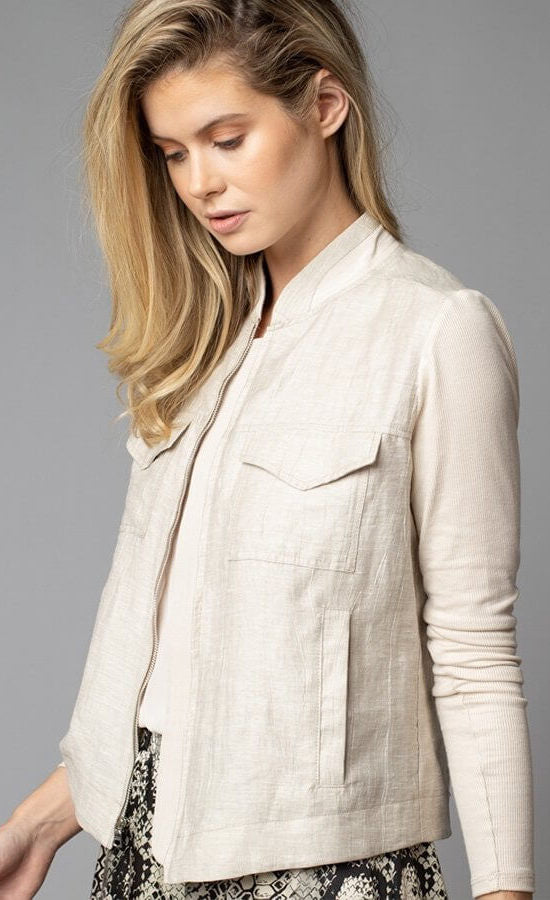 Front, top half left side view of a woman wearing the Lola & Sophie Crinkle Linen Jean Jacket. The sand colored jacket features two front, fold over breast pockets, a zipper front, a linen body, and fitted long sleeves.