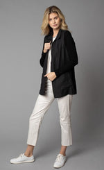 Load image into Gallery viewer, Front, left-sided full body view of a woman wearing white capris and the Lola &amp; Sophie Crinkle Linen Zip-Front Long Black Jacket. This jacket has a flat collar, a zip up front, front pockets, and fitted long sleeves.
