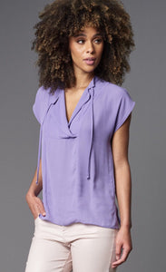 Front top half view of a woman wearing the lola & sophie Double Georgette Cap-Sleeve Top. this top is lavender colored and has a v-neck with drawstrings that can be tied together.