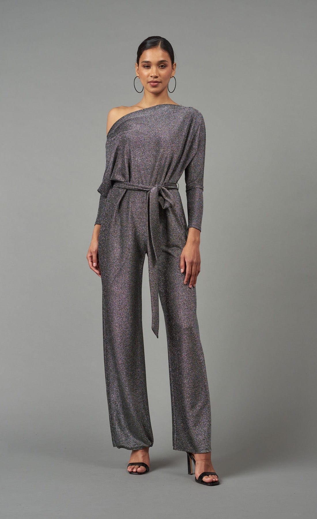 Front full body view of the lola & sophie lurex jersey jumpsuit. This jumpsuit is silver colored with long sleeves, long wide pants, a tie belt at the waist, and a boat neck being worn off the shoulder.