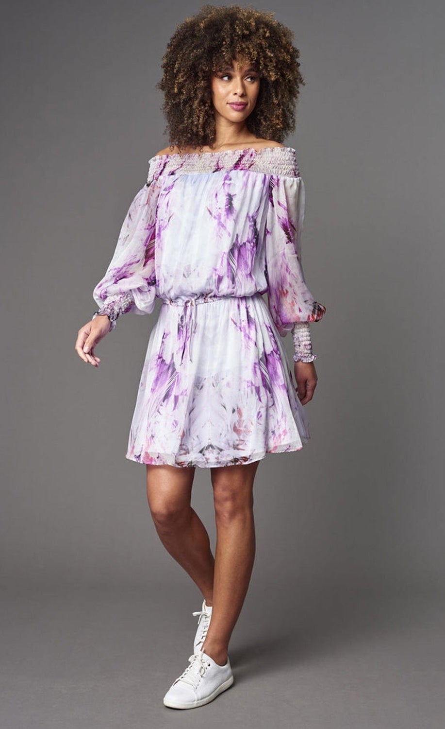 Front full body view of a woman wearing the lola & sophie Painted Flower Chiffon Off-Shoulder Dress. This dress is white with lilac/pink painted flowers. The dress has bishop sleeves with elastic cuffs and a drawstring waistband.