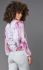 Load image into Gallery viewer, Back top half view of a woman wearing the lola &amp; sophie painted flower top. This top is white with a pink and red painted design sporadically on it. This top has long sleeves, a keyhole back, and a drawstring hem.
