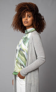 Left side, top-half view of a woman with her hands in the pocket of her white pants. The woman is also wearing green printed tank under the lola & sophie split back cardi. This cardigan is grey and draped open. The long sleeves are fitted and ribbed.