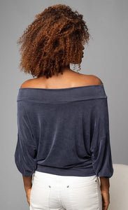 Back, top half view of a woman wearing white pants and the Lola & Sophie Off the Shoulder Top. This top is a greyish-midnight blue. The sleeves are rolled up to the elbows.