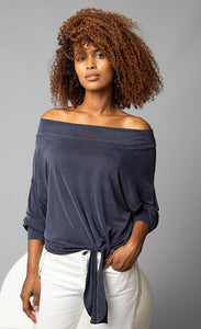 Front, top half view of a woman with her left hand in the pockets of her white jeans. On the top she is wearing the Lola & Sophie Off the Shoulder Top. This top is a greyish-midnight blue. The sleeves are rolled up to the elbows and the front has a left sided tie at the bottom.