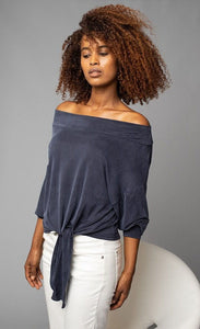 Front, top half view of a woman wearing white pants and the Lola & Sophie Off the Shoulder Top. This top is a greyish-midnight blue. The sleeves are rolled up to the elbows and the front has a left sided tie at the bottom.