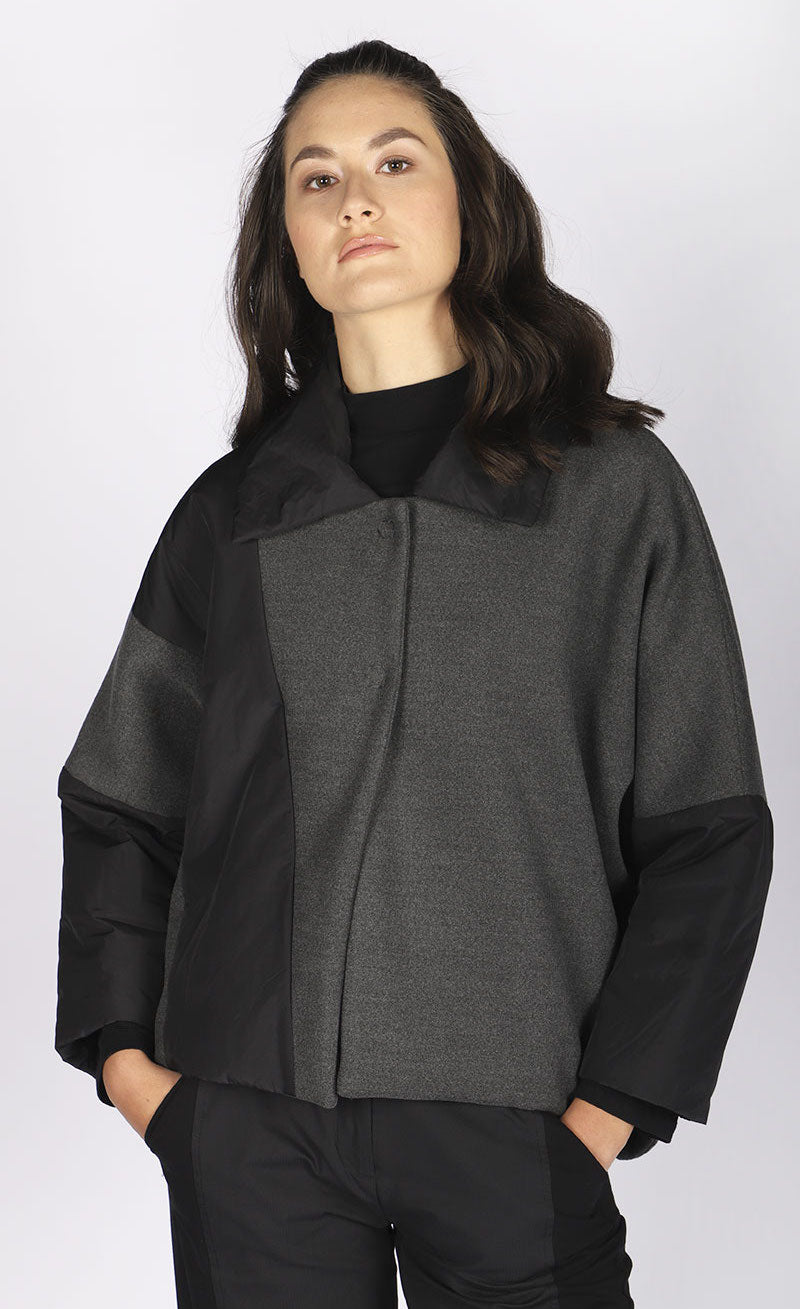 Front top half view of a woman wearing the luukaa short anthracite coat. This coat has a hidden front closure, and an anthracite body with black puffer fabric on the flat collar and long sleeves. The black puffer fabric also runs diagonally across the body