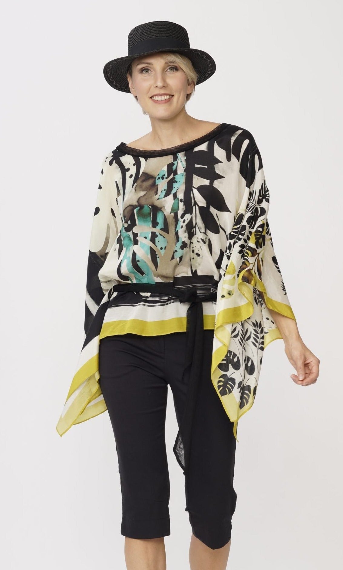 front top half view of a model wearing the luukaa belted poncho. This poncho is white with mustard trim and black and teal leaf print all over it. 