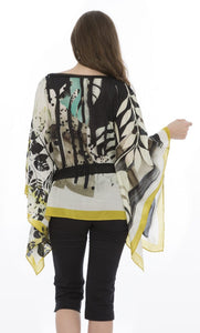 Back top half view of a model wearing the luukaa belted poncho. This poncho is white with mustard trim and black and teal leaf print all over it.