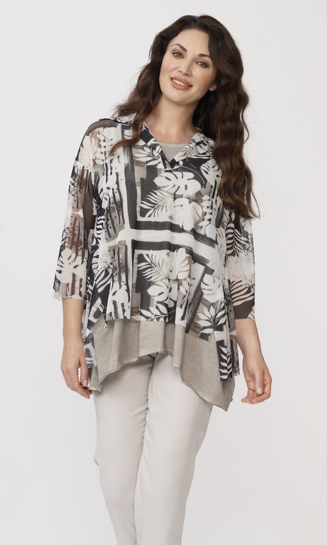 Front top half view of a woman wearing white capris and the luukaa leaf print top over a grey tank. This top is sheer with white and grey leaf print. The sleeves are 3/4 length, the neck is a v-neck, and the hem is short in the front and long on the sides.