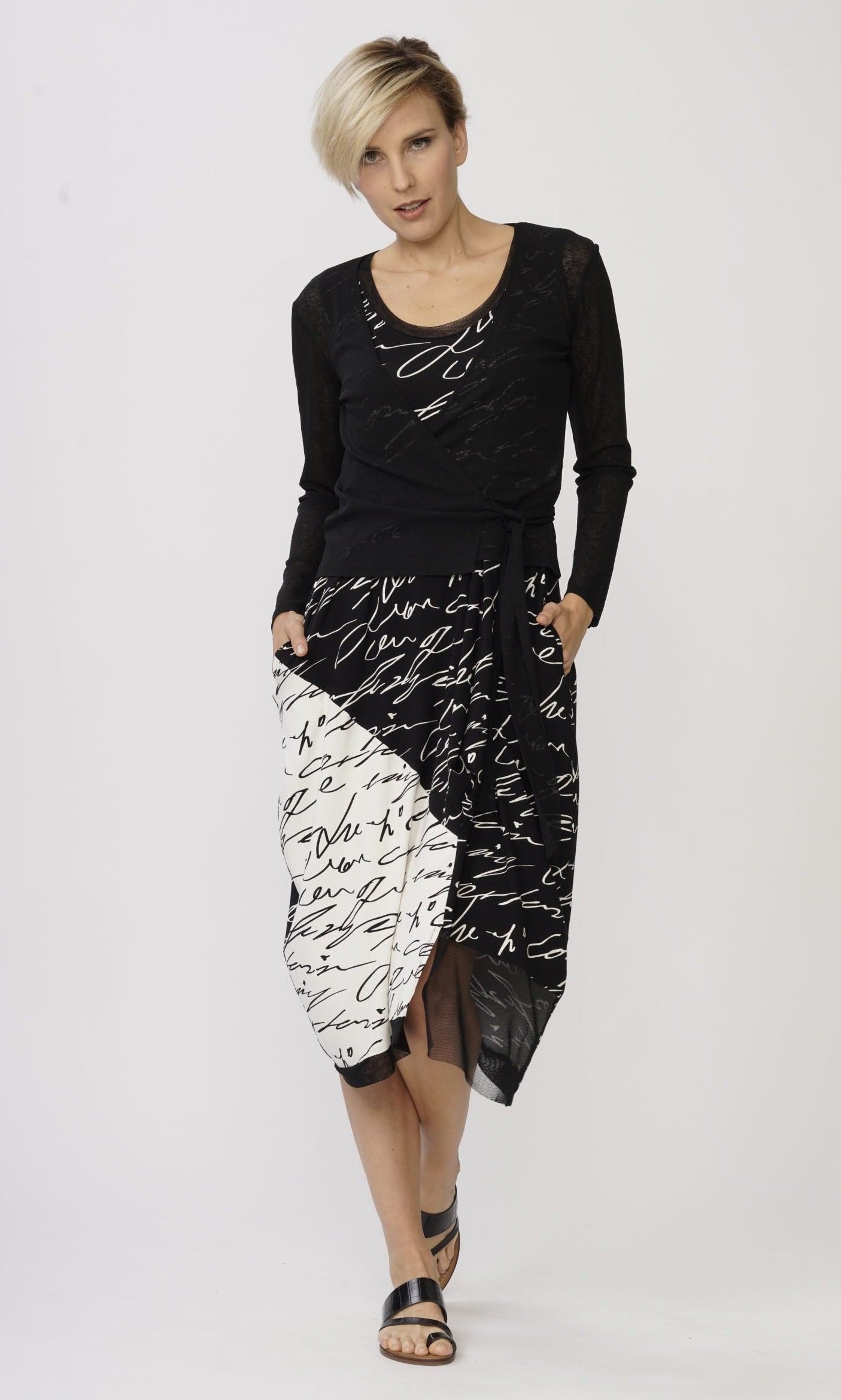 Front full body view of a woman wearing the luukaa scribbles dress. The dress is covered with a short black wrap jacket. The dress is black with white handwriting. The right side near the bottom has white with black handwriting. The dress comes down below the knees with some mesh trim on the right side of the bottom hem.