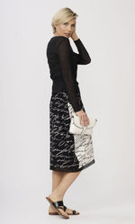 Load image into Gallery viewer, Right side full body view of a woman wearing the luukaa scribbles dress. The dress is covered with a short black wrap jacket. The dress is black with white handwriting on the front and back. The right side near the bottom has white with black handwriting. The dress comes down below the knees with some mesh trim on the hem.
