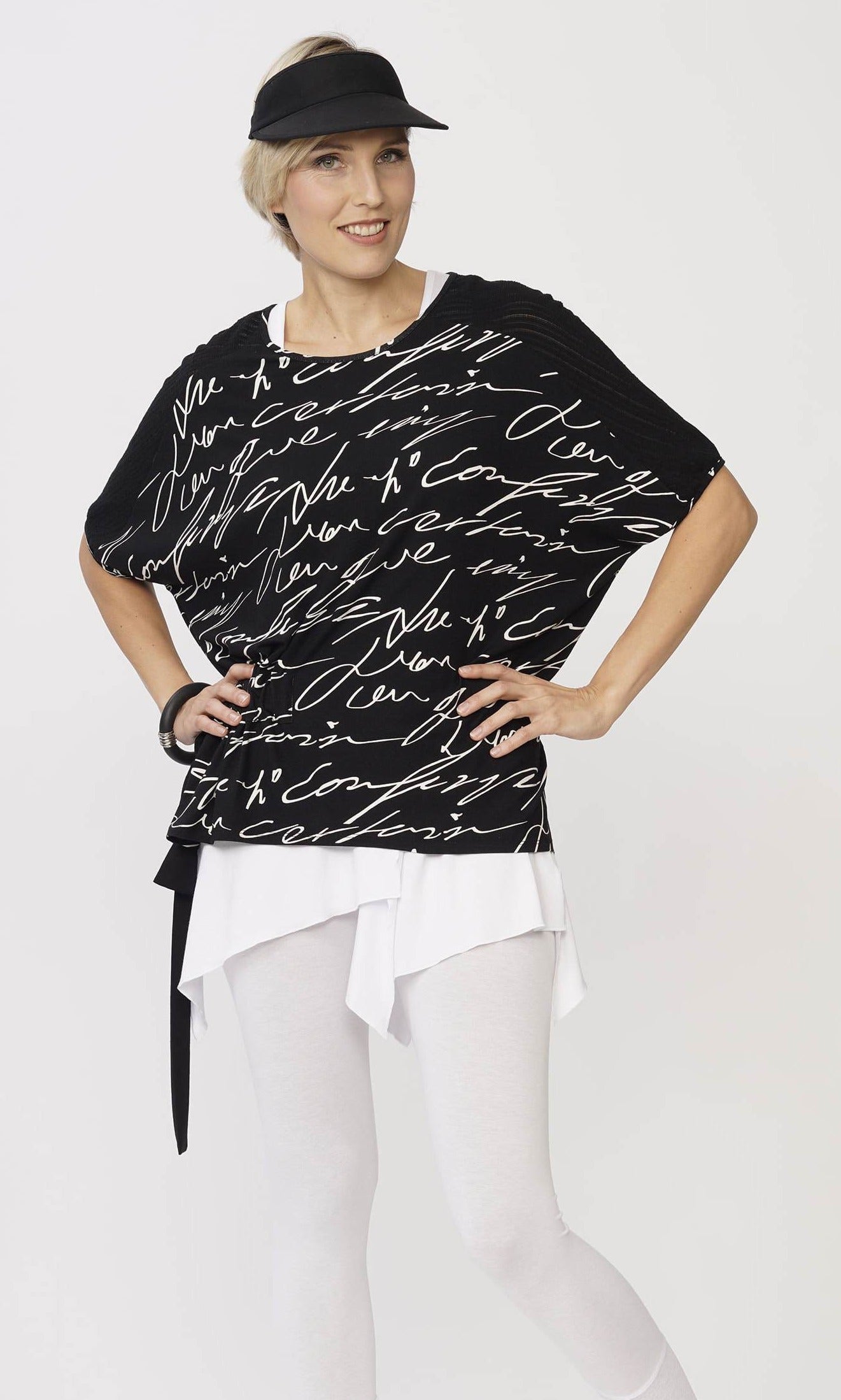 Top half, front view of a woman wearing the luukaa scribble top in black. This black top has white handwriting all over it, a side adjustable belt that can be tied, and elbow length dolman sleeves.