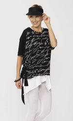 Load image into Gallery viewer, Top half, front view of a woman wearing the luukaa scribble top in black. This black top has white handwriting all over it, a side adjustable belt that can be tied, and elbow length dolman sleeves.
