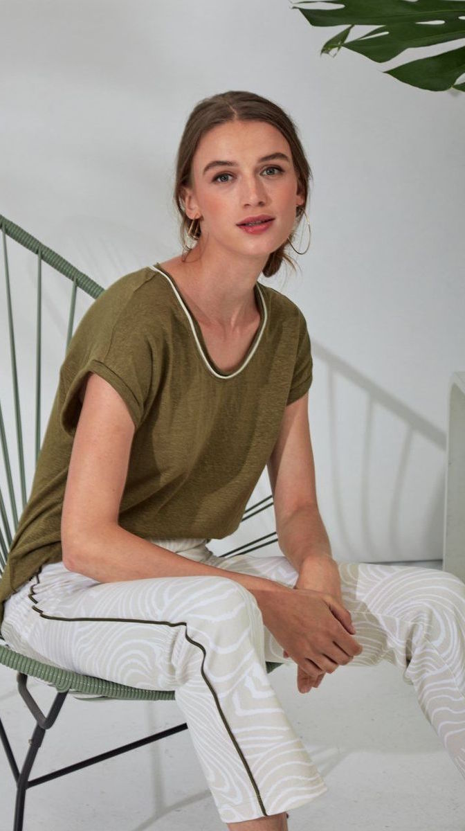 Front view of a woman sitting and wearing white pants and the lysse classic tee. this tee is jungle khaki colored. It has short drop shoulder sleeves, a high low hem, and white edging around the round neck. 