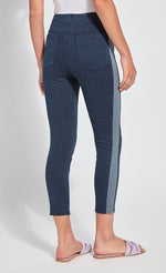 Load image into Gallery viewer, Back view of the bottom half of a woman wearing the Lysse Nomad Crop Leggings. These leggings are dark denim with a side blue and white houndstooth printed stripe going down the entire leg. The leggings have two large back pockets and cut off above the ankles. The woman is also wearing flat beige sandals.
