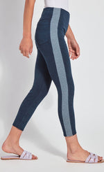 Load image into Gallery viewer, Right side view of the bottom half of a woman with one leg in front of the other and wearing the Lysse Nomad Crop Leggings. These leggings are dark denim with a side blue and white houndstooth printed stripe going down the entire leg. The leggings cut off above the ankles and the woman is wearing flat beige sandals.
