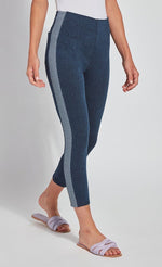 Load image into Gallery viewer, Front, right side view of the bottom half of a woman wearing the Lysse Nomad Crop Leggings. These leggings are dark denim with a side blue and white houndstooth printed stripe going down the entire leg. The leggings cut off above the ankles and the woman is wearing flat beige sandals.
