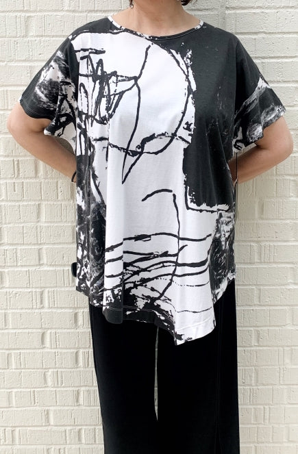 Front top half view of a woman wearing the moyuru short sleeve printed top. This top is white with black abstract print and an asymmetrical hem.