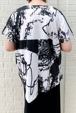Load image into Gallery viewer, Back top half view of a woman wearing the moyuru short sleeve printed top. This top is white with black abstract print and an asymmetrical hem.
