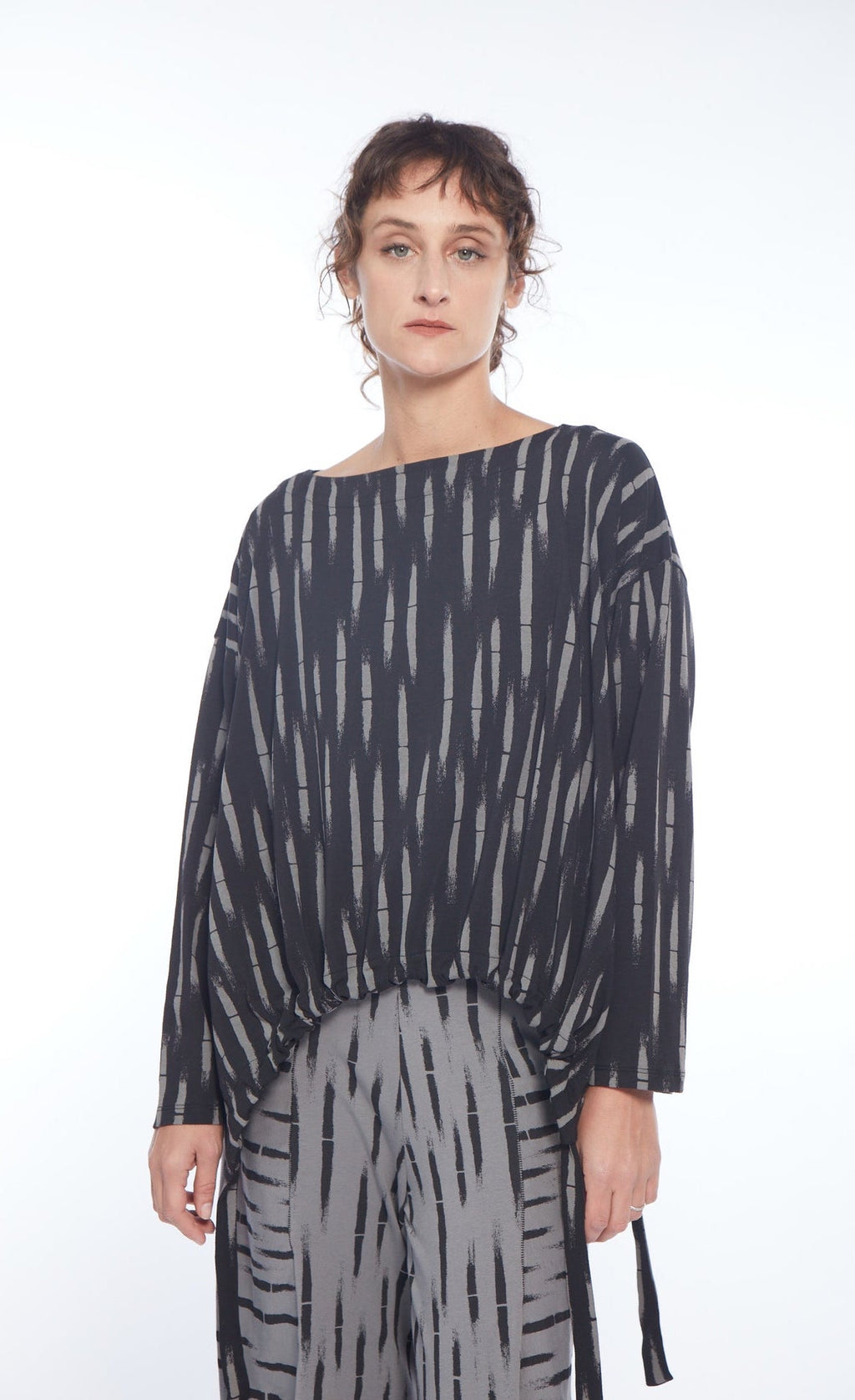 Front top half view of a woman wearing the mxmatthildur liev top. This top is black with a grey bamboo print on it. The top has long sleeves, a boat neck, and a tie/elastic hem.