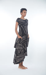 Load image into Gallery viewer, Front, right side full body view of a woman wearing the matthidul safari muse dress. This dress is black with an abstract taupe print on it. The dress has cap sleeves, front pockets, and an asymmetrical hem.
