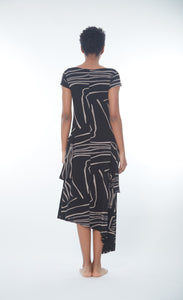 Back full body view of a woman wearing the matthidul safari muse dress. This dress is black with an abstract taupe print on it. The dress has cap sleeves and an asymmetrical hem.