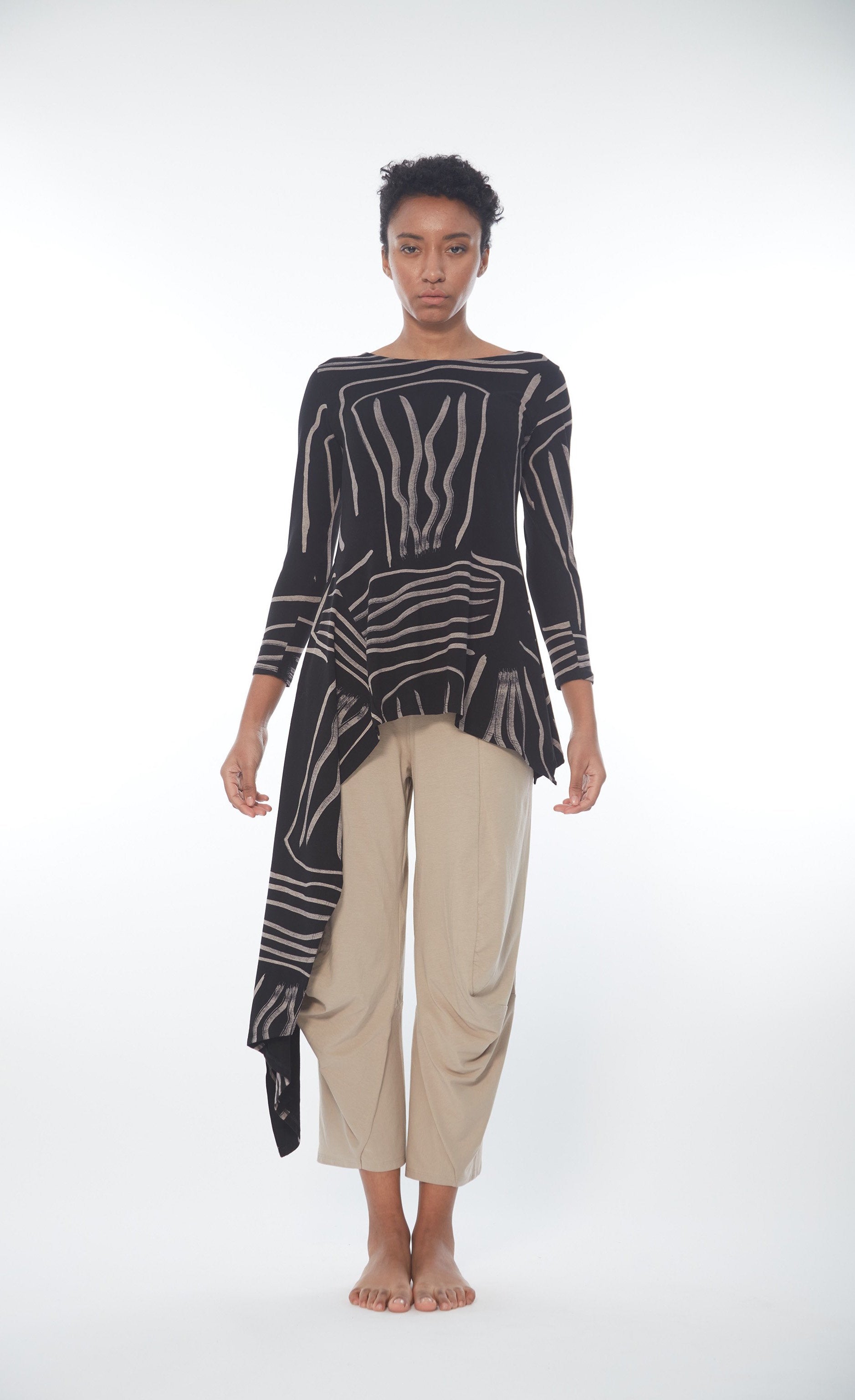 Front full body view of a woman wearing the Matthildur Safari Deck top. This top is black with an abstract taupe print on it. The shirt has 3/4 length sleeves, a boat neck, and a longer right side that goes down below the knees.
