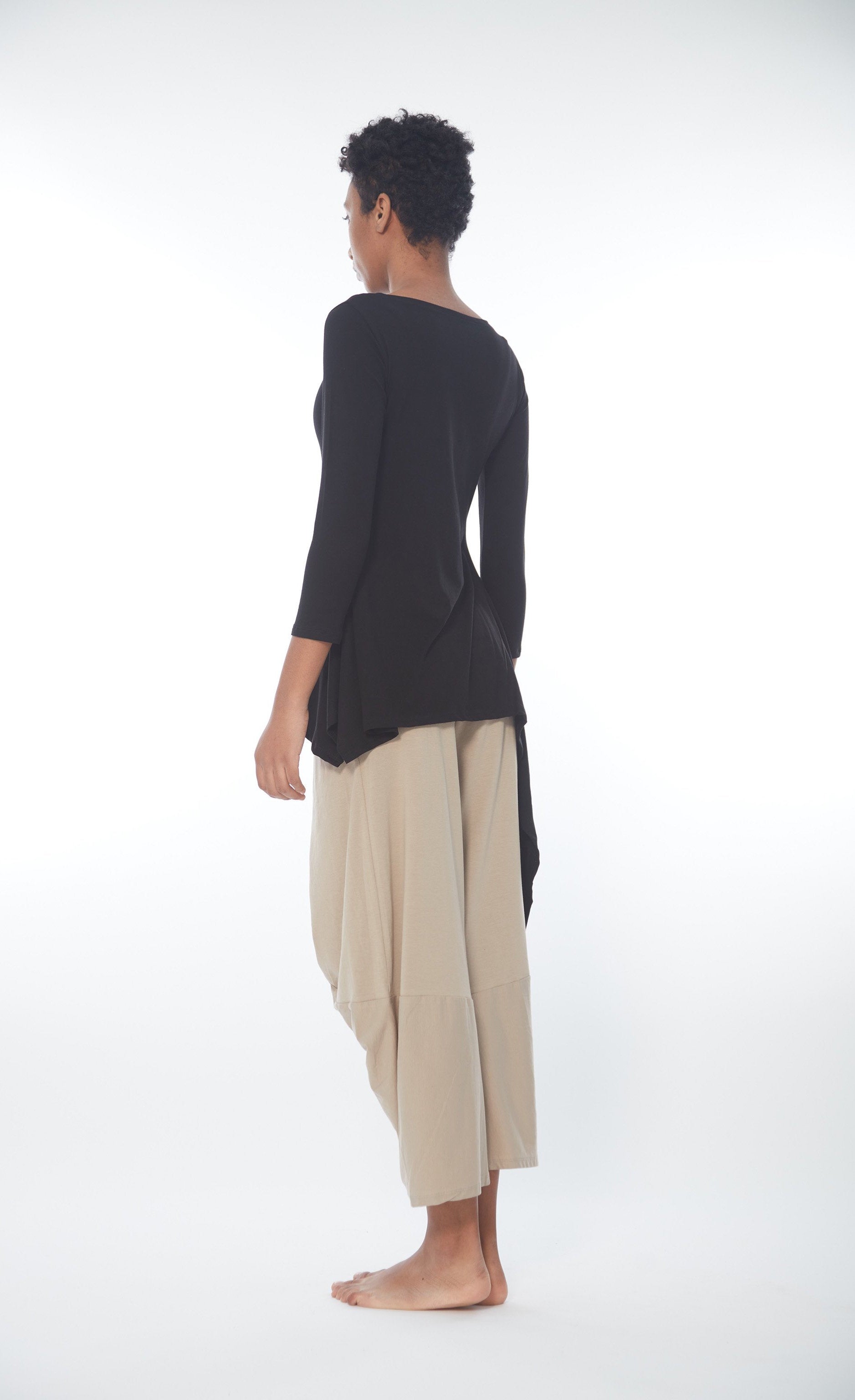 Back full body view of a woman wearing the Matthildur Safari Deck top. This image shows the black version of this top. The shirt has 3/4 length sleeves, a boat neck, and a longer right side that goes down below the knees.