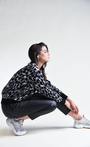 Right side full body view of a woman crouching down and wearing black pants and the molly bracken animal print sweater. This sweater has grey, black, and with animal print with black trim on the cuffs, hem, and round neckline.