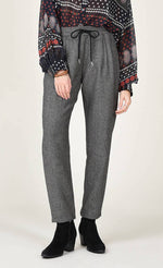 Load image into Gallery viewer, Front bottom half view of a woman wearing the molly bracken tzigane print shirt and the molly bracken grey herringbone pant. This pant has an elastic waistband with a black tie and a wider fit with legs that taper in at the bottom.
