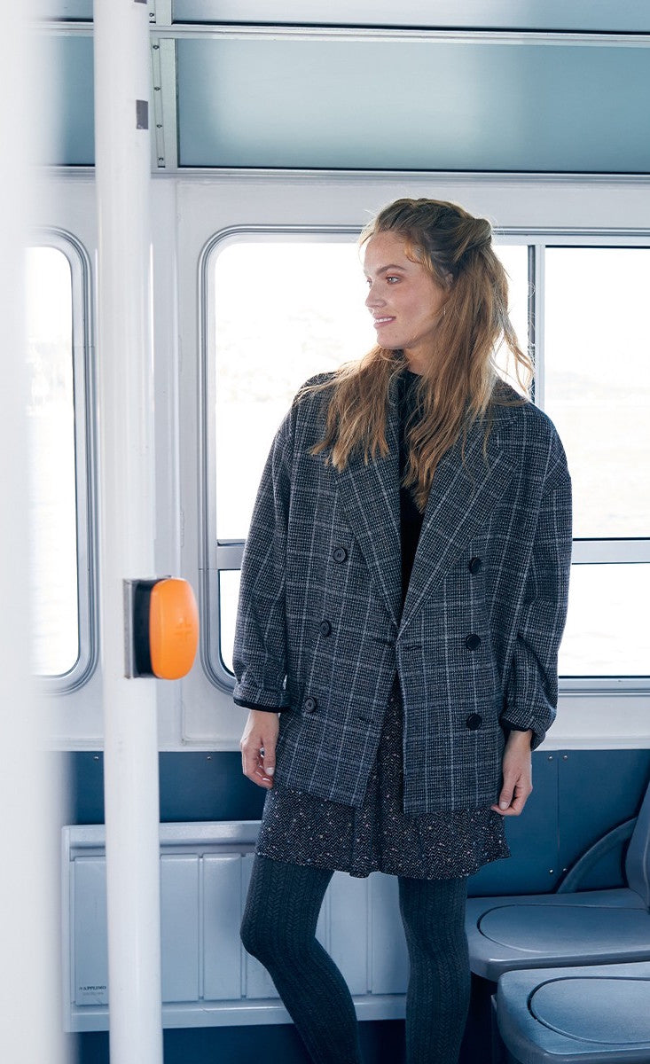 Front full body view of a woman wearing the molly bracken plaid caban coat and a skirt with tights. This oversized coat is grey with double breasted buttons and a large collar.