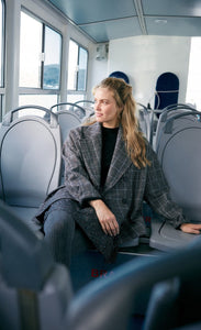 Front full body view of a woman sitting on a bus wearing the molly bracken plaid caban coat and a skirt with tights. This oversized coat is grey with double breasted buttons and a large collar.