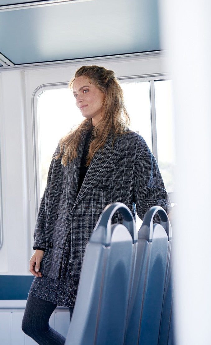 Front full body view of a woman wearing the molly bracken plaid caban coat and a skirt with tights. This oversized coat is grey with double breasted buttons and a large collar.