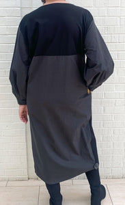 Back full body view of a woman wearing the moyuru black dress. This dress hits below the knees and has a straight silhouette, oversized long sleeves made of a structured fabric, and a mix of fabrics on the back