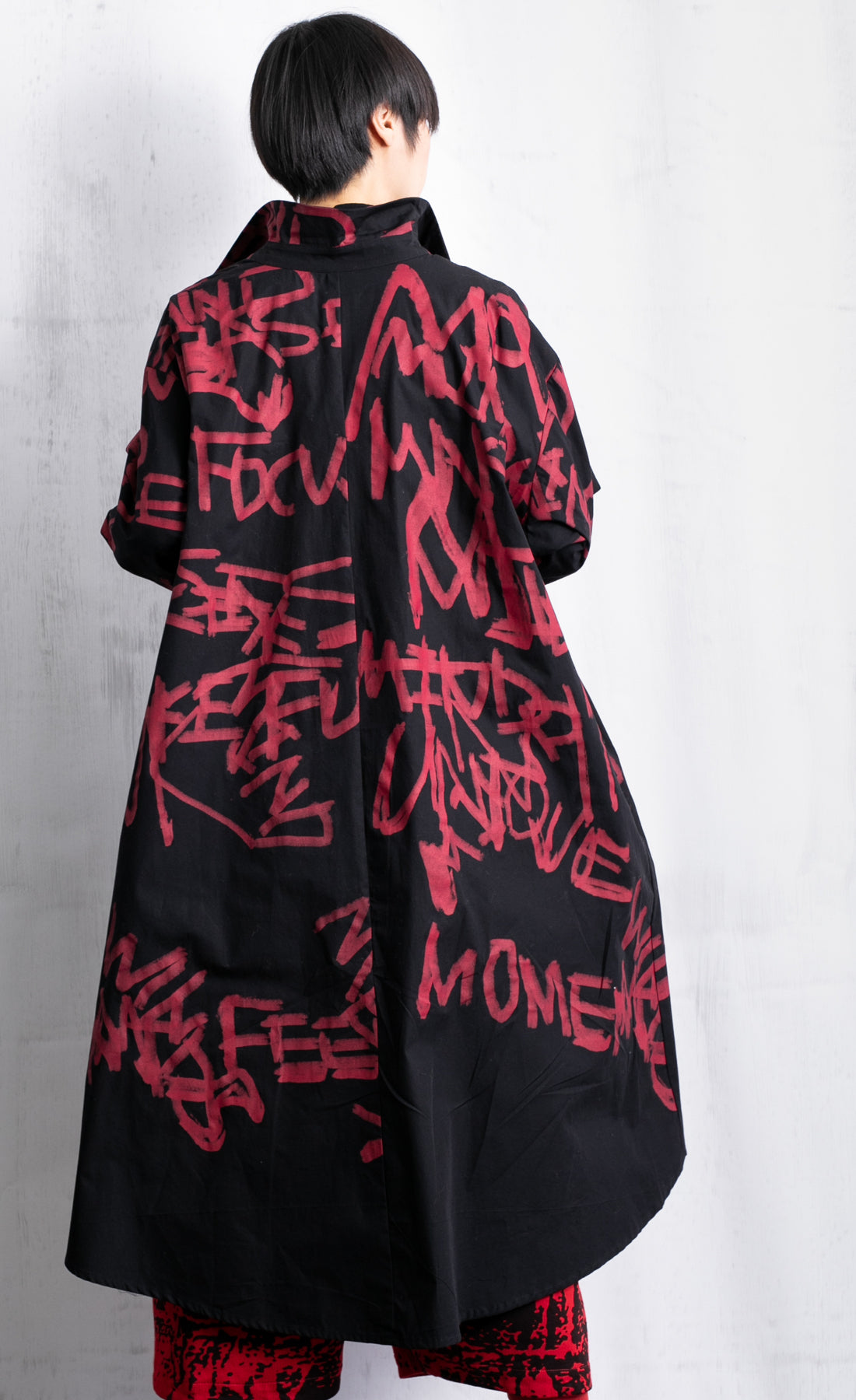 Back full body view of a woman wearing red pants and a black shirt/dress with red scribble print all over it. This shirt dress has a button down front, long sleeves, and comes down to the knees.