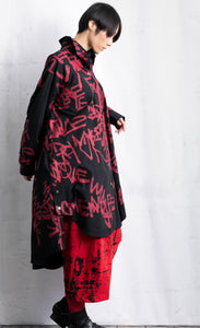 Right side full body view of a woman wearing red pants and a black shirt/dress with red scribble print all over it. This shirt dress has a button down front, long sleeves, and comes down to the knees.