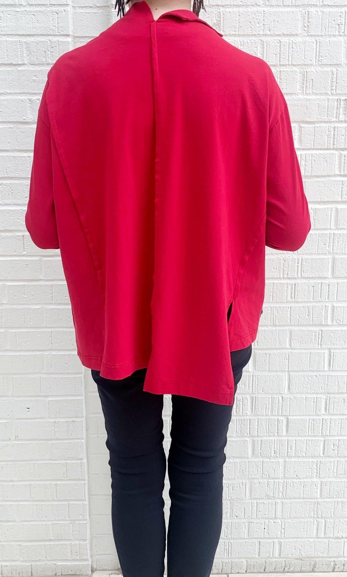 Back full body view of a woman wearing black pants and the moyuru red pullover top. This top has long sleeves, a boxy fit, and an asymmetrical hem.