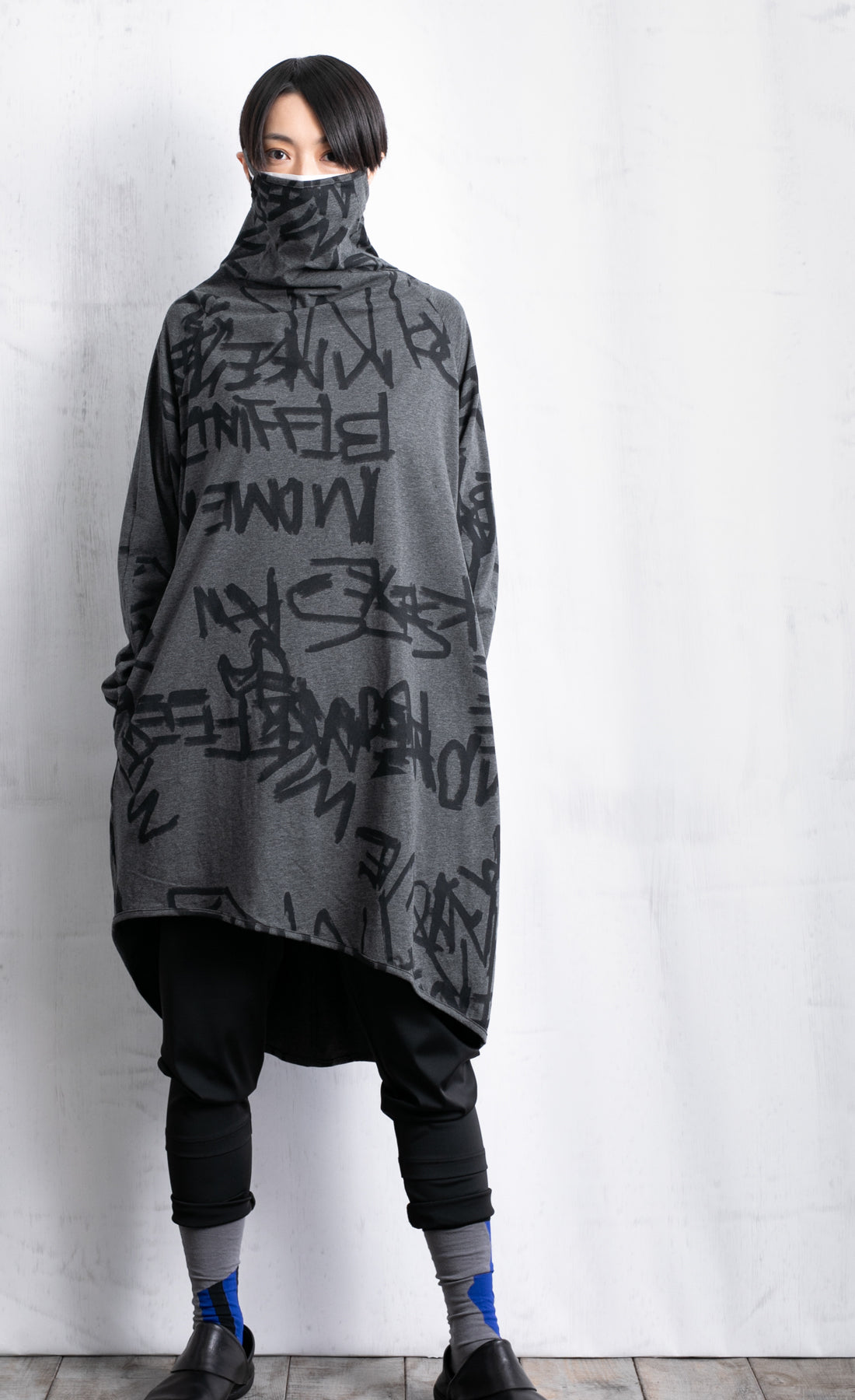 Front full body view of a woman wearing black pants and the moyuru charcoal scribble tunic. This tunic is dark grey with a graffiti/scribble print all over it. The Tunic has long sleeves and a cowl neck that is being used as a face covering.