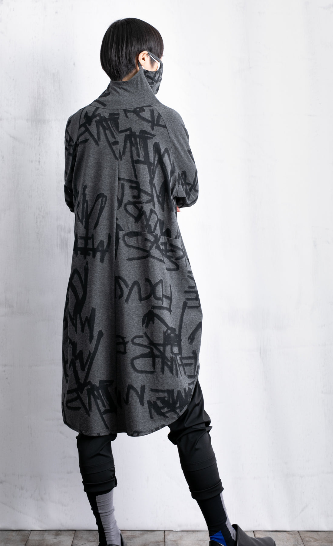 Back full body view of a woman wearing black pants and the moyuru charcoal scribble tunic. This tunic is dark grey with a graffiti/scribble print all over it. The Tunic has long sleeves and a cowl neck that is being used as a face covering.