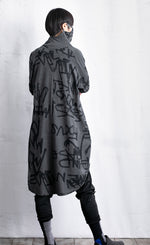 Load image into Gallery viewer, Back full body view of a woman wearing black pants and the moyuru charcoal scribble tunic. This tunic is dark grey with a graffiti/scribble print all over it. The Tunic has long sleeves and a cowl neck that is being used as a face covering.
