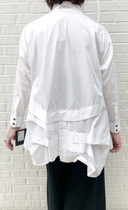 Back full body view of a woman wearing the moyuru white shirt. This shirt has an asymmetrical hem on the back with tucks and pleats and a center slit. The shirt also has long sleeves.