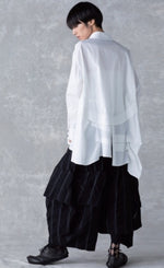 Load image into Gallery viewer, Back full body view of a woman wearing the moyuru white shirt. This shirt has an asymmetrical hem on the back with tucks and pleats and a center slit. The shirt also has long sleeves.
