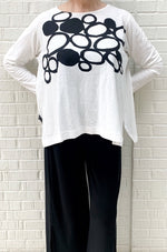 Load image into Gallery viewer, Front top half view of a woman wearing the moyuru long sleeve circle top. This top is white with black filled and empty circles on the top half. The top has fitted sleeves and a flowy body.
